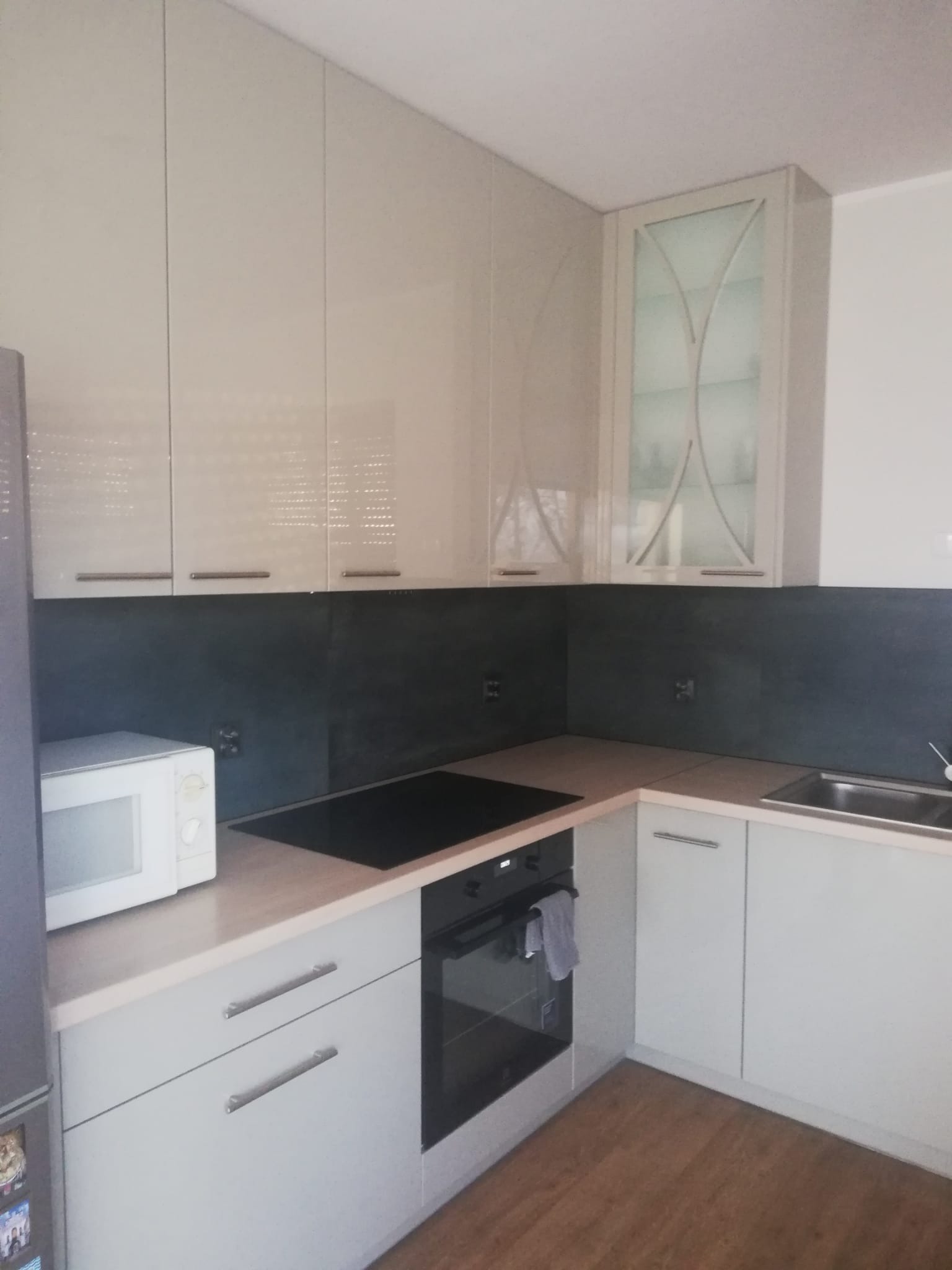 Jasna kuchnia z błyszczącymi frontami szafek, czarnym blatem i sprzętem AGD. Górne szafki z przeszkleniami. Ujęcie z perspektywy użytkownika.