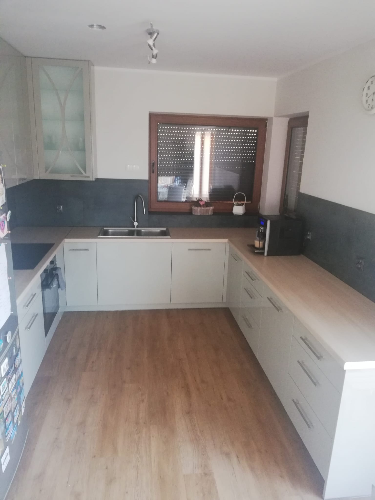 Jasna kuchnia z drewnianą podłogą i oknem. Na blacie ekspres do kawy. Górne szafki z witrynami. Ciemna ściana nad blatem. Minimalistyczny styl.