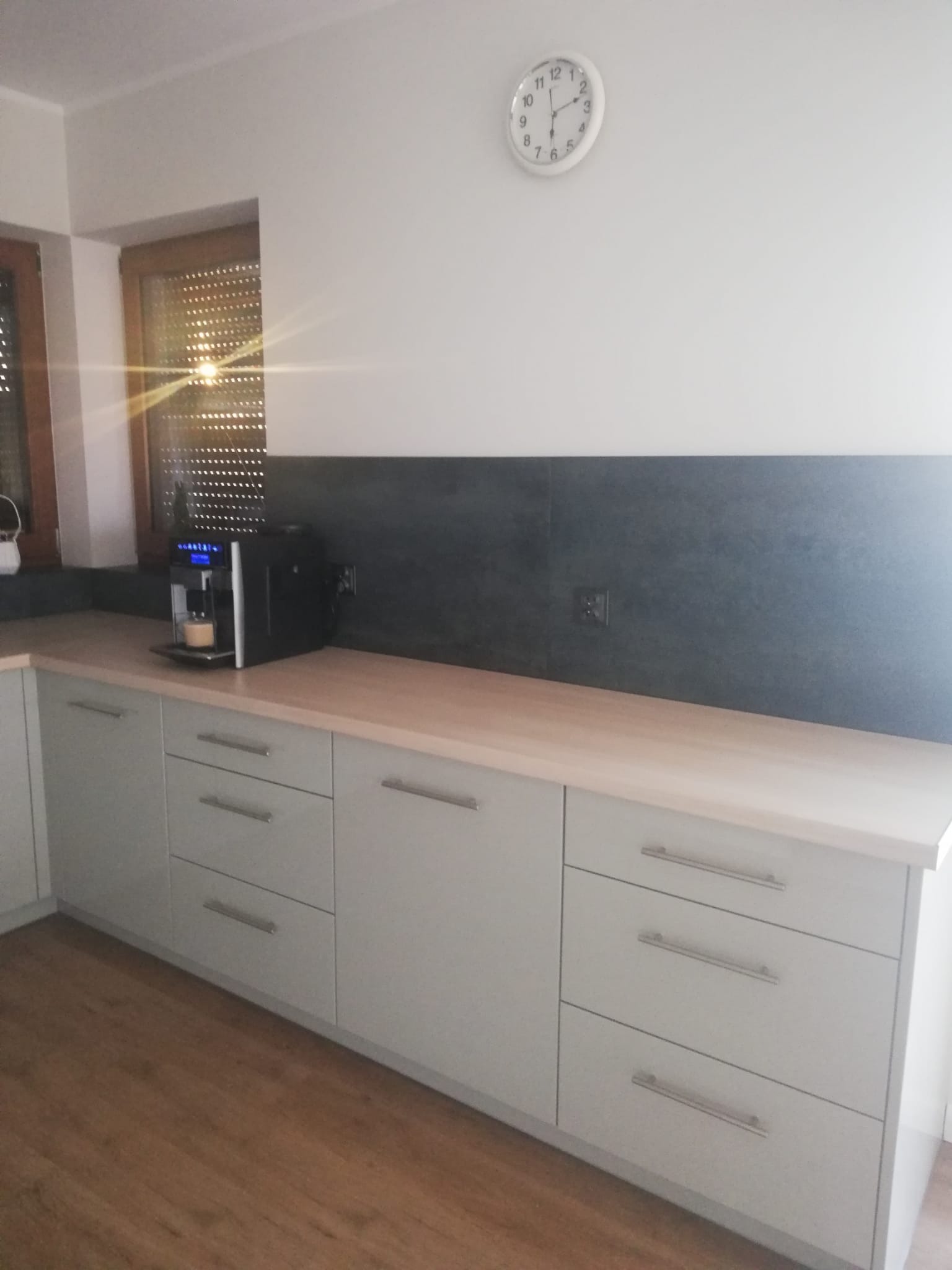 Jasnoszara zabudowa kuchenna z drewnianym blatem i czarnym panelem ściennym. Na blacie stoi ekspres do kawy. Na ścianie wisi biały zegar.