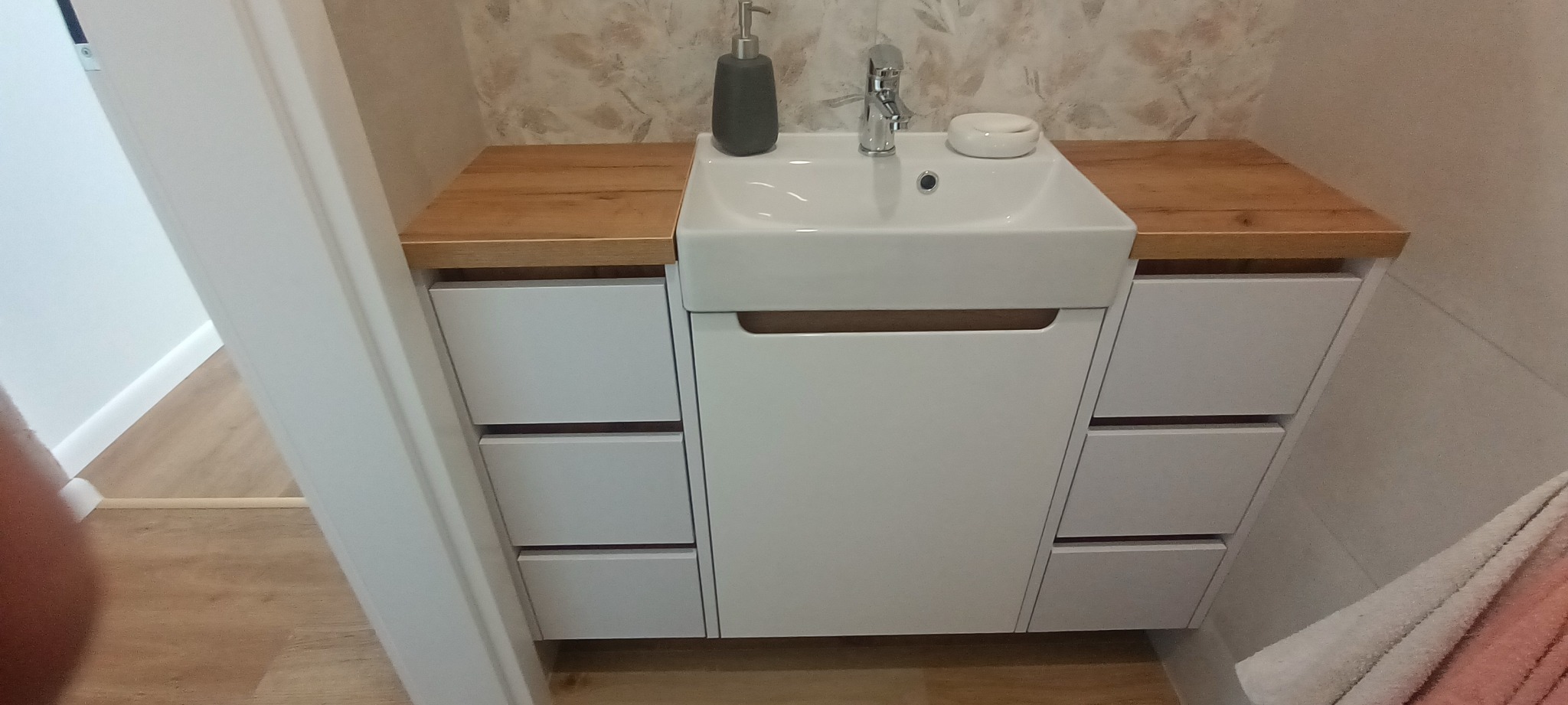 Nowoczesna szafka łazienkowa z drewnianym blatem, białe fronty z minimalistycznymi uchwytami, umywalka nablatowa i armatura.