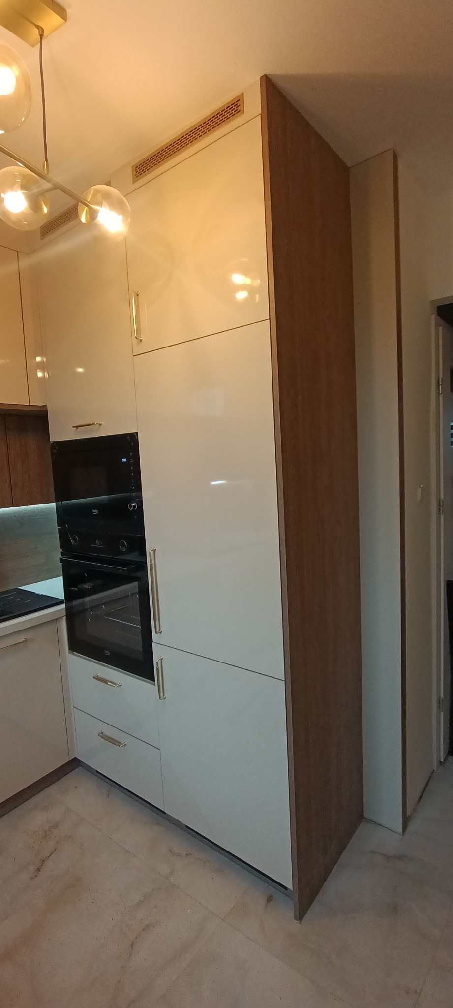 Biała zabudowa kuchenna z piekarnikiem w nowoczesnym stylu, złote uchwyty, drewniane wykończenie boku szafki, widoczna kratka wentylacyjna pod sufitem.