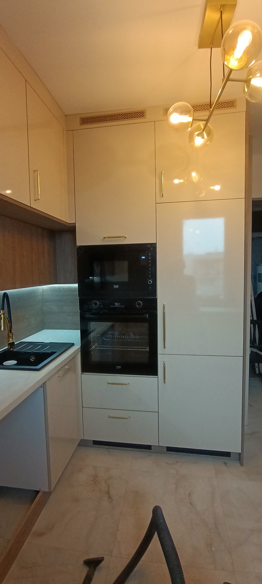 Beżowa kuchnia z zabudową piekarnika i mikrofalówki Beko, czarny zlew z baterią w kolorze złota, oświetlenie sufitowe. Widok na fragment podłogi i odkurzacz.