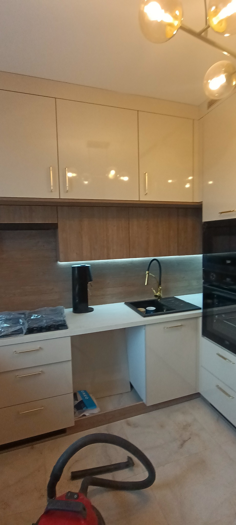 Jasna kuchnia z błyszczącymi frontami i drewnianym panelem. Na blacie czarny dystrybutor wody i zlewozmywak z złotą baterią. Widoczny odkurzacz na podłodze.