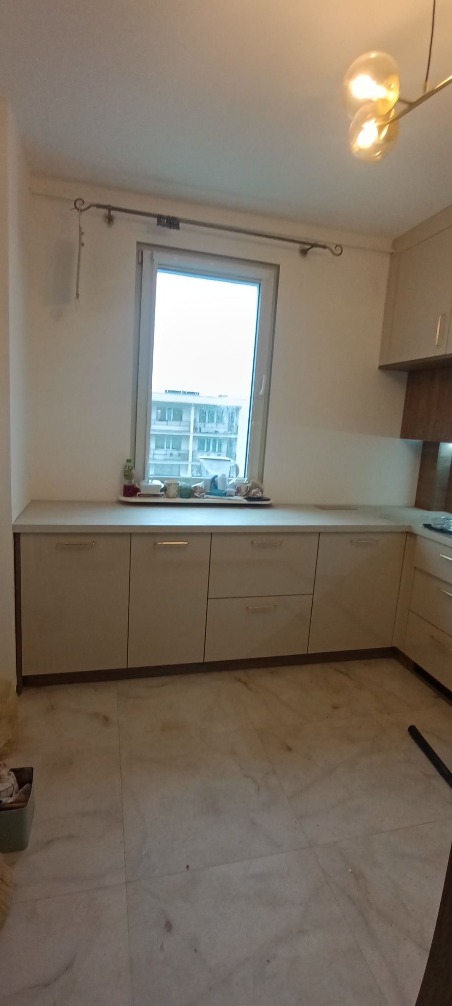 Jasna kuchnia z beżowymi frontami szafek, marmurową podłogą i blatem pod oknem. Na blacie widoczne naczynia i akcesoria kuchenne. Widok na budynek przez okno.