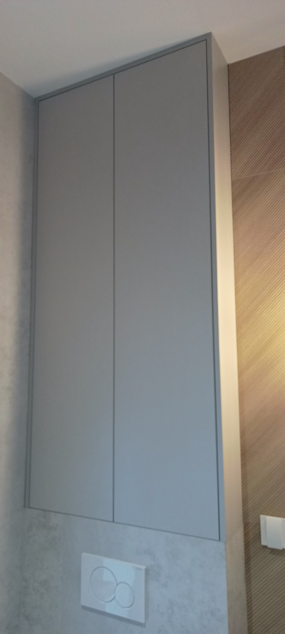 Szara szafka wisząca z dwoma frontami, zamontowana nad podtynkową spłuczką WC w łazience z mozaikową ścianą i teksturowaną tapetą.