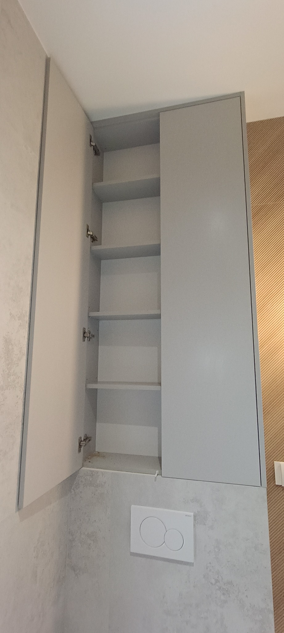 Otwarta, szara szafka wisząca z półkami w łazience w Lubawie. Minimalistyczny design, widoczne zawiasy i przycisk spłuczki.