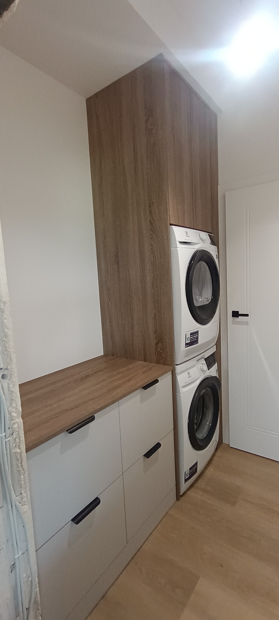 Zabudowa z szafek z blatem drewnopodobnym i szafki wiszącej w odcieniu drewna, wkomponowana w nią pralka i suszarka automatyczna.