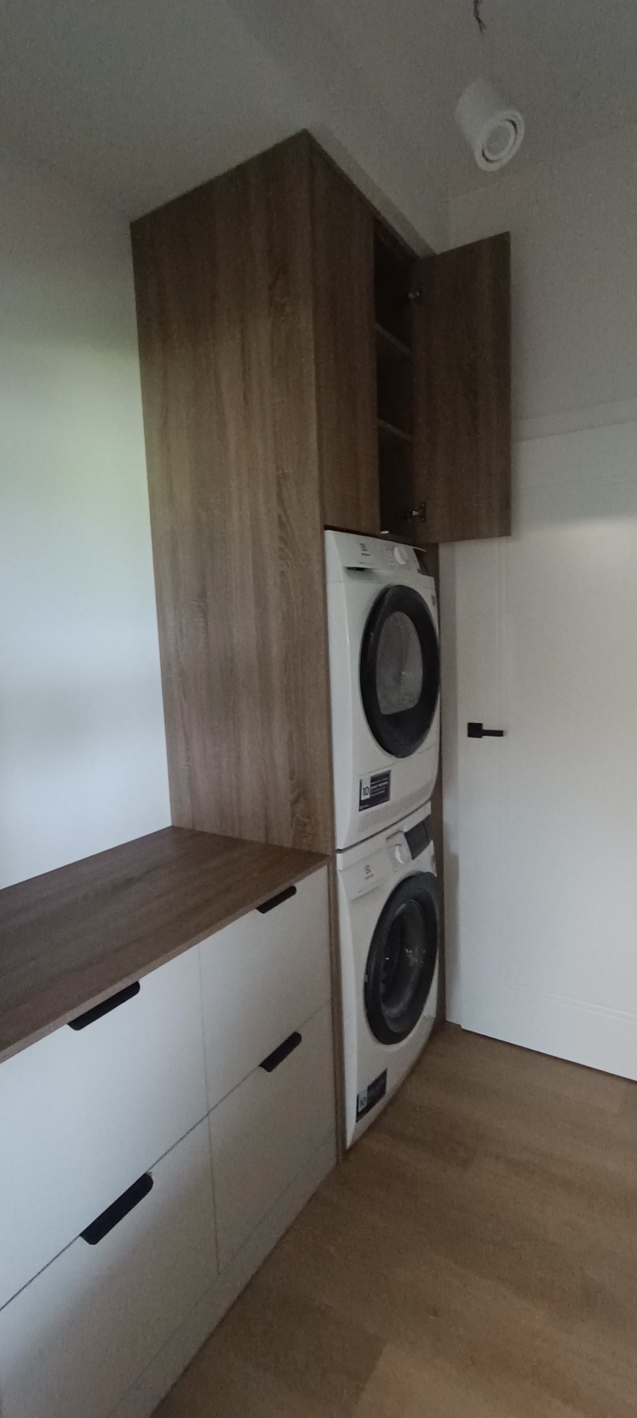 Zabudowa pralki i suszarki z szafkami na wymiar, fronty białe z czarnymi uchwytami, drewnopodobny blat i górne szafki, jedna szafka otwarta.