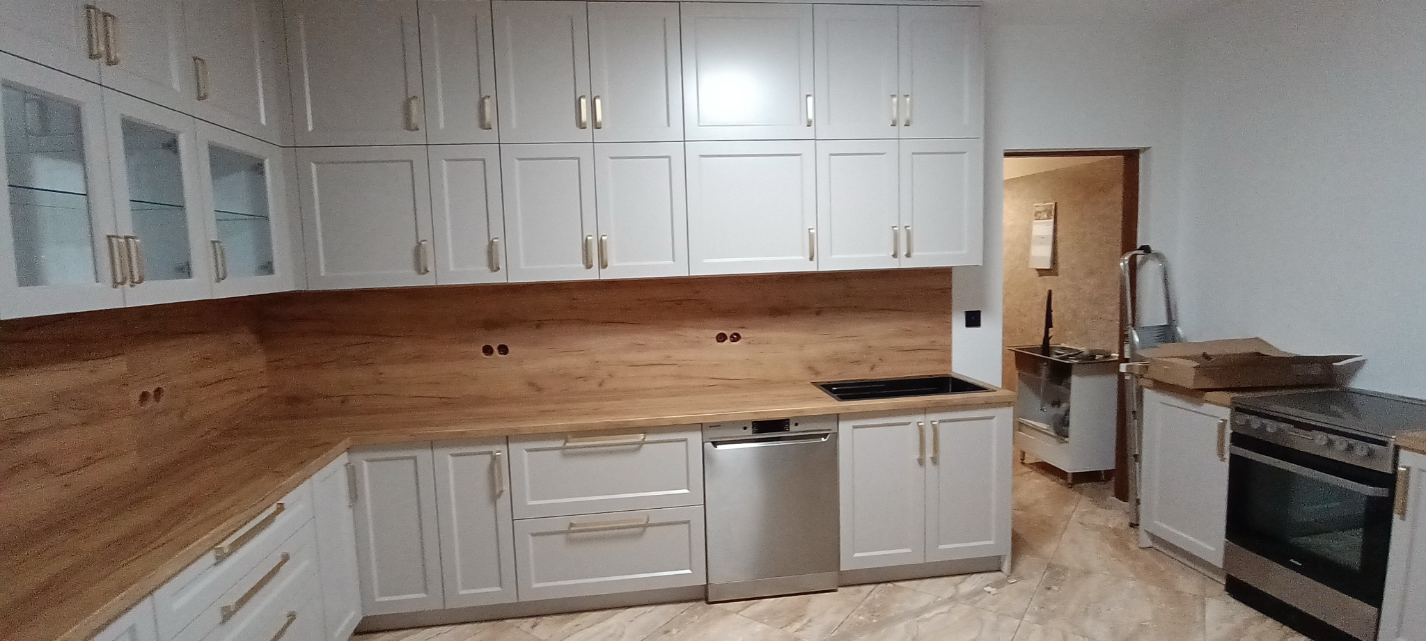 Jasna kuchnia z drewnianym blatem i złotymi uchwytami, widok na zlew i sprzęt AGD. Częściowo widoczny sprzęt w przejściu do innego pomieszczenia.