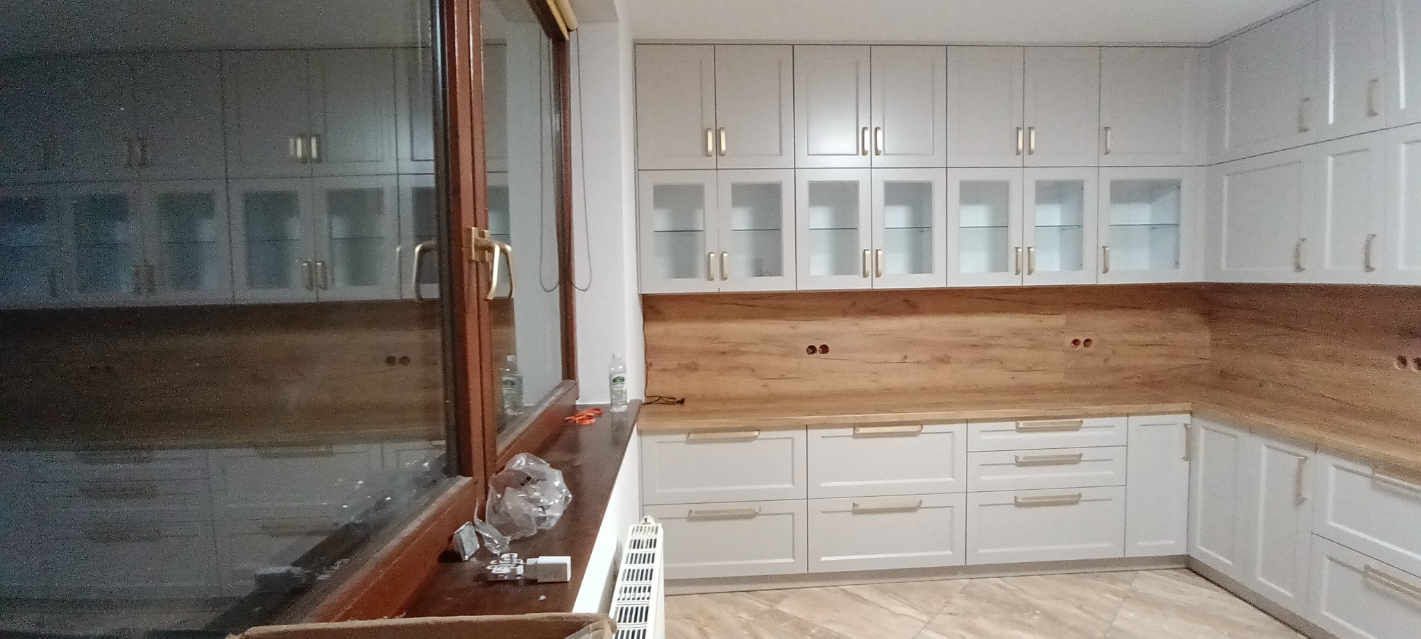 Jasna kuchnia na wymiar z drewnianym blatem i przeszklonymi szafkami wiszącymi. Widoczne gniazdka elektryczne w ścianie nad blatem. Ujęcie z perspektywy okna.