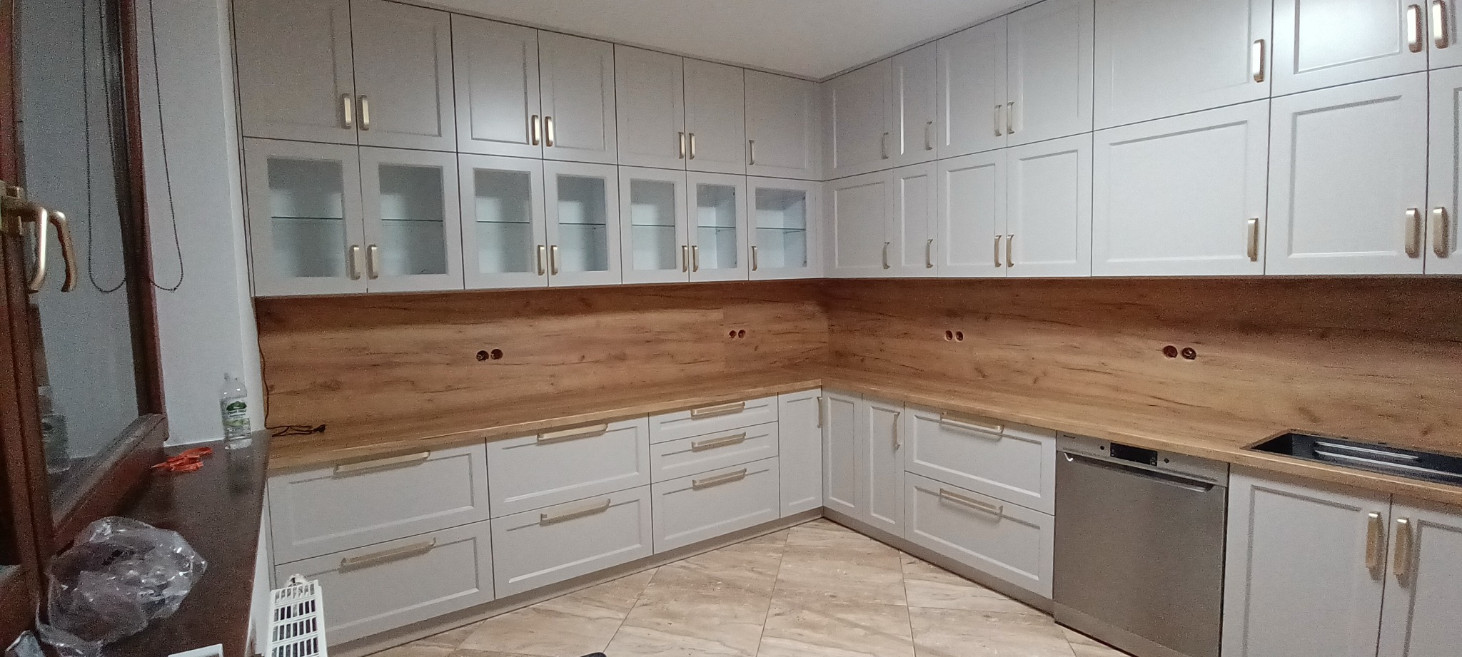 Jasnoszara zabudowa kuchenna z drewnianym blatem i złotymi uchwytami. Górne szafki częściowo przeszklone. Widoczne gniazdka elektryczne w ścianie.