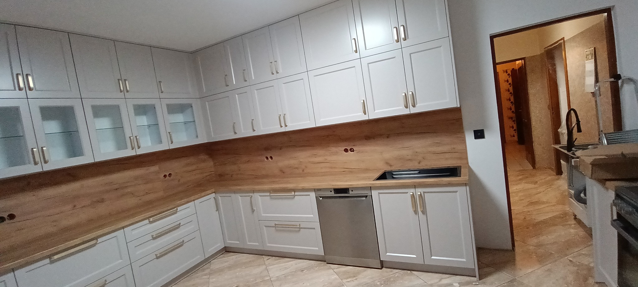 Jasna kuchnia z drewnianym blatem i białymi szafkami, widok na wnętrze przez otwarte drzwi. Nowoczesny design, złote uchwyty, płyta grzewcza w blacie.