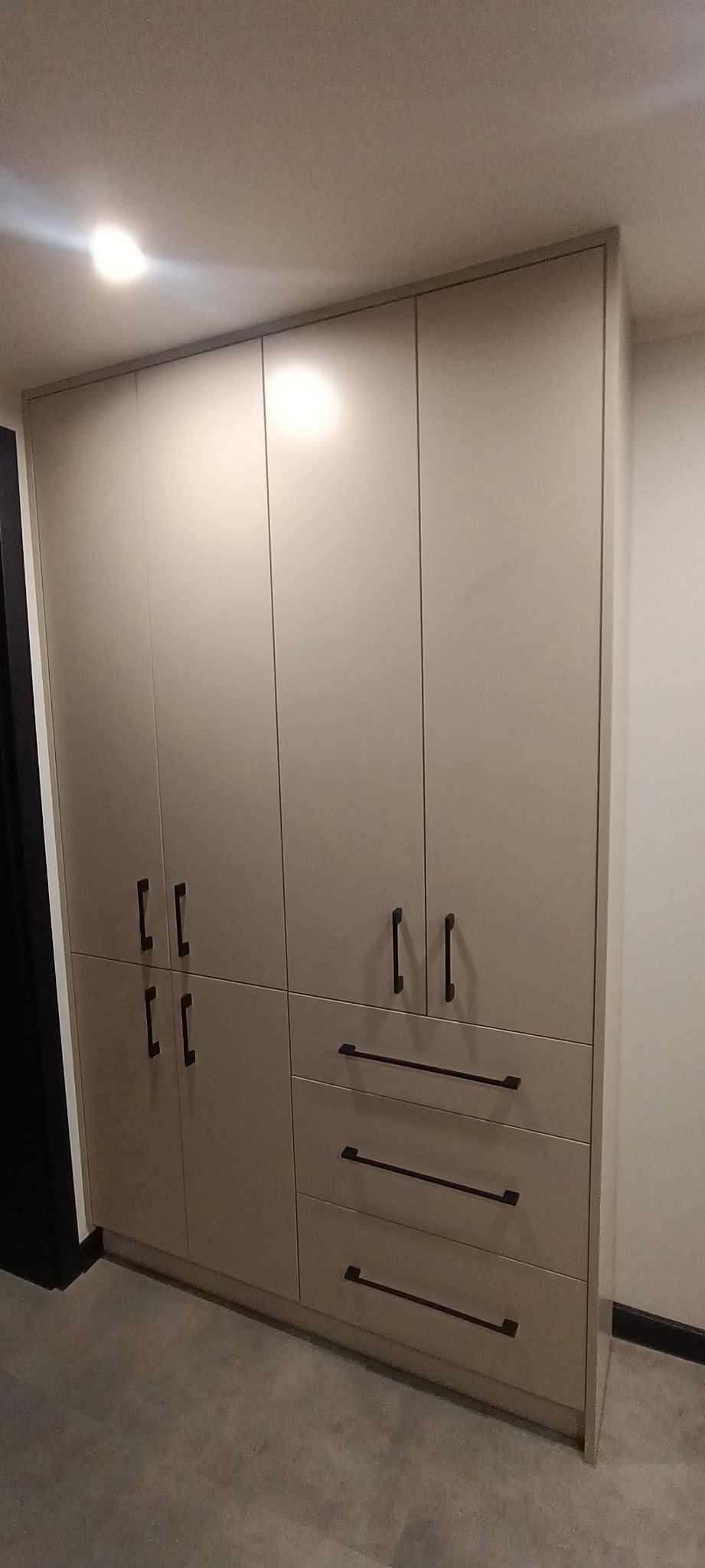 Jasna szafa wnękowa z ciemnymi, podłużnymi uchwytami, podzielona na segmenty z drzwiczkami i szufladami. Minimalistyczny design, neutralne tło.