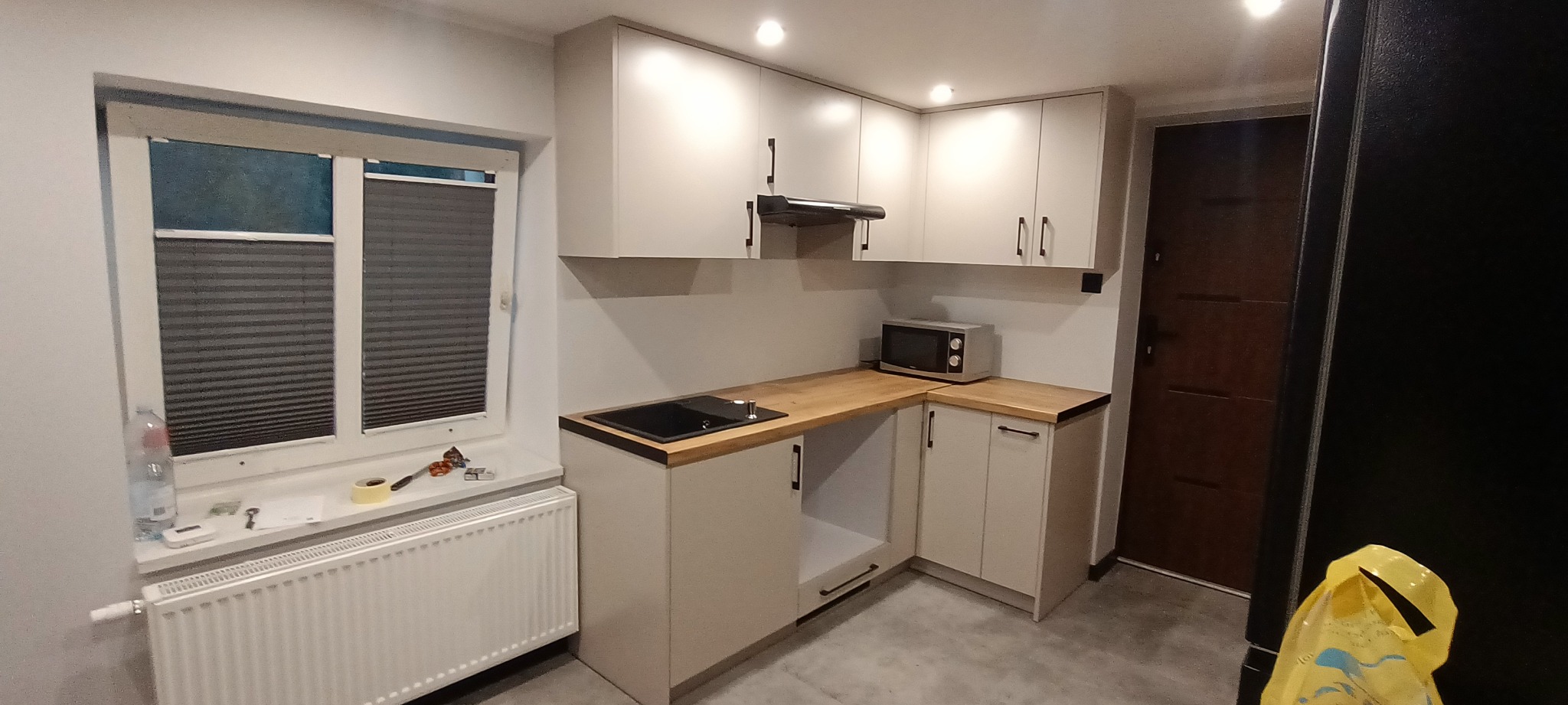 Jasna kuchnia z drewnianym blatem i czarną płytą grzewczą. Nad blatem wiszące szafki z czarnymi uchwytami. Na blacie mikrofalówka. Przy oknie grzejnik.