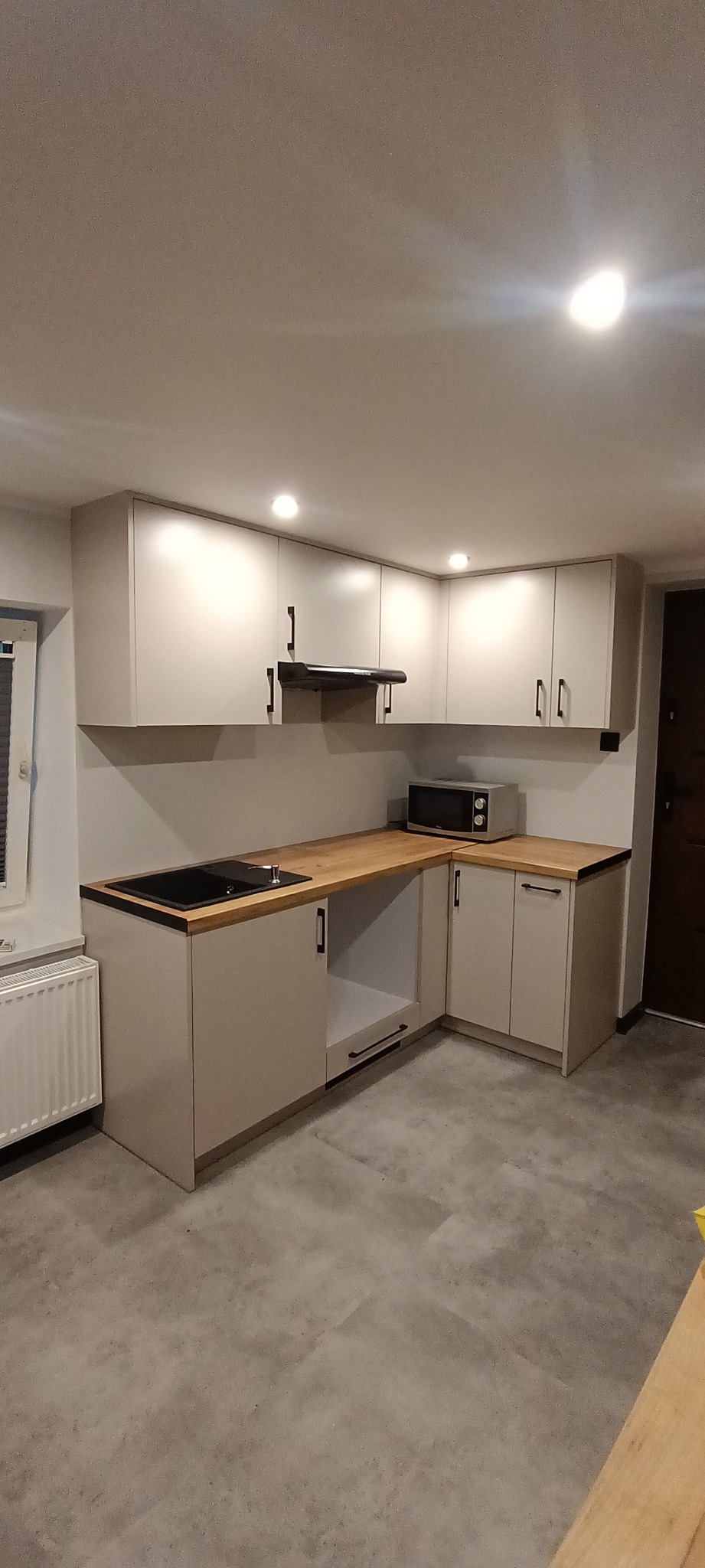 Jasna, narożna kuchnia z drewnianym blatem i czarnymi uchwytami. Nad blatem wiszące szafki z oświetleniem punktowym. W rogu mikrofalówka. Minimalistyczny design.