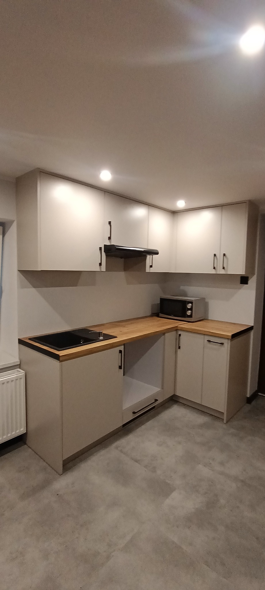 Jasna, narożna zabudowa kuchenna z drewnianym blatem i czarnymi uchwytami. Widoczny zlew, okap i mikrofalówka. Minimalistyczny design.