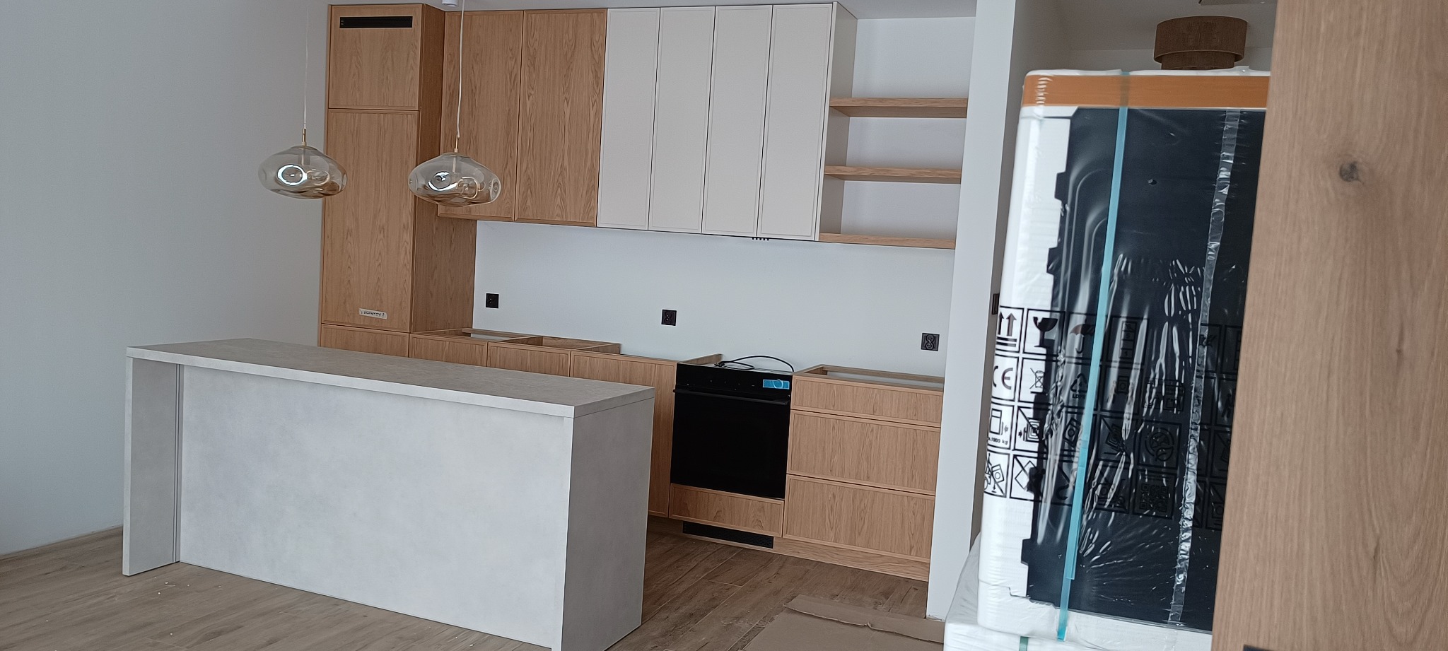 Jasna kuchnia z drewnianymi i białymi szafkami, wyspą i lampami wiszącymi. Widoczny fragment zapakowanego sprzętu AGD.