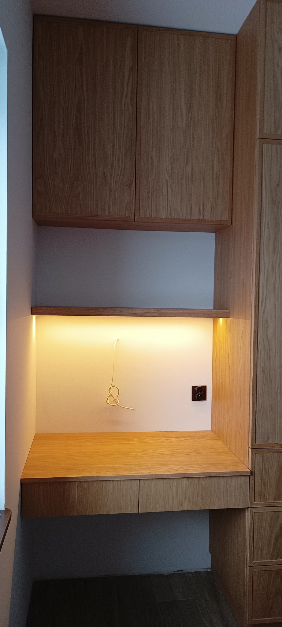 Drewniany, zabudowany kącik biurowy z oświetleniem LED pod półką i gniazdkiem elektrycznym. Widoczne szafki wiszące i szuflady. Naturalne wykończenie drewna.