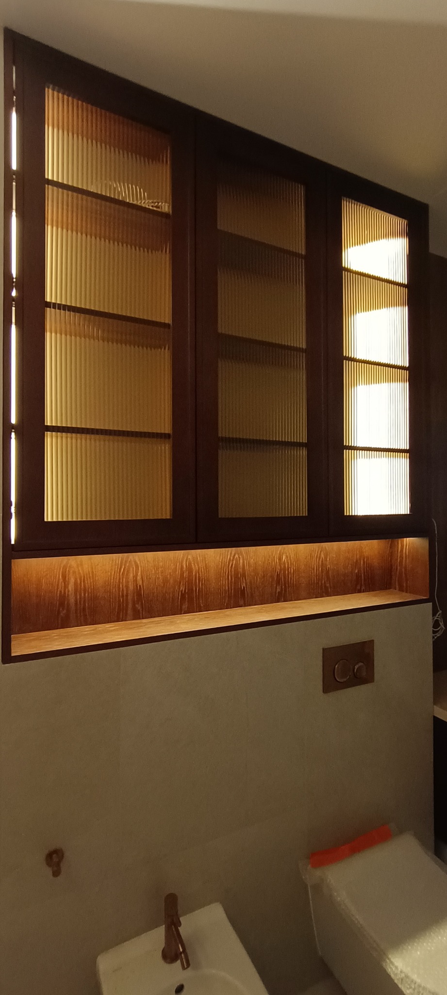 Zabudowa meblowa z ryflowanymi szybami i podświetleniem LED nad umywalką. Ciemne drewno, ciepłe światło, nowoczesny design. Widoczne detale armatury.