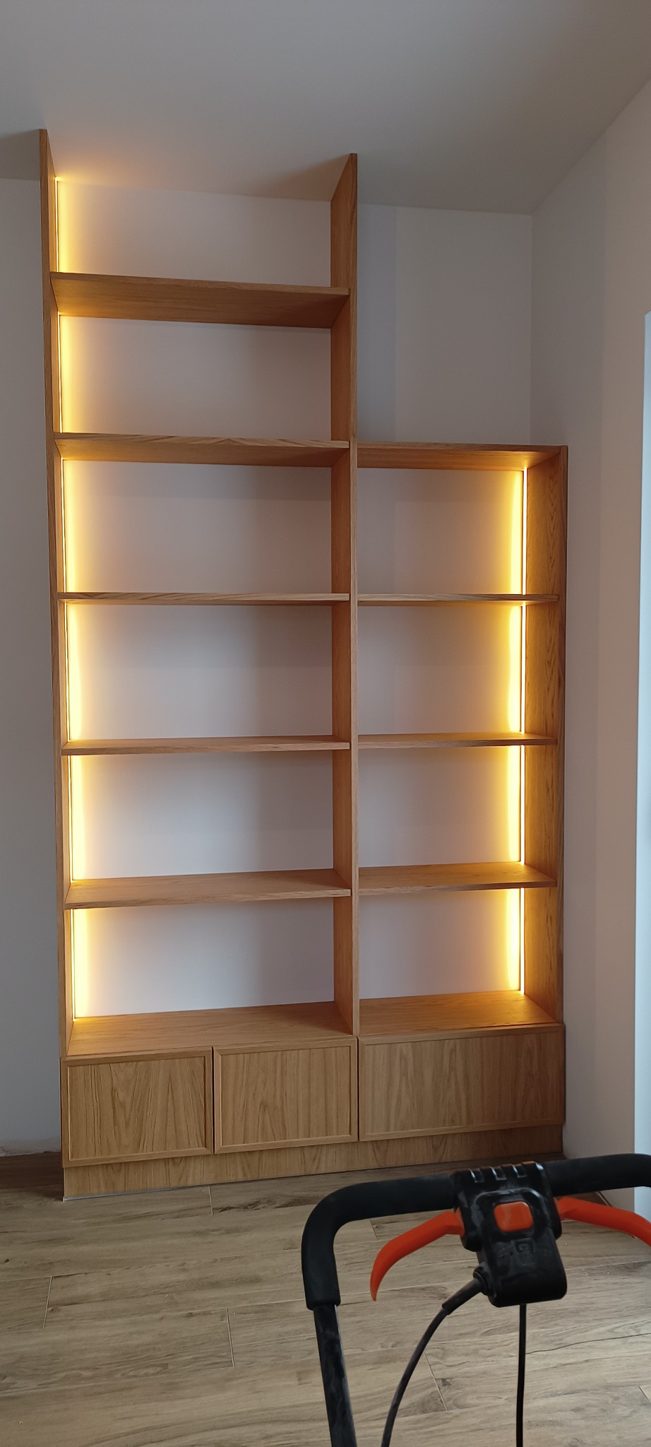 Drewniana szafa wnękowa z oświetleniem LED, widok z dołu. Naturalne wykończenie drewna, minimalistyczny design. Na podłodze widoczny odkurzacz.