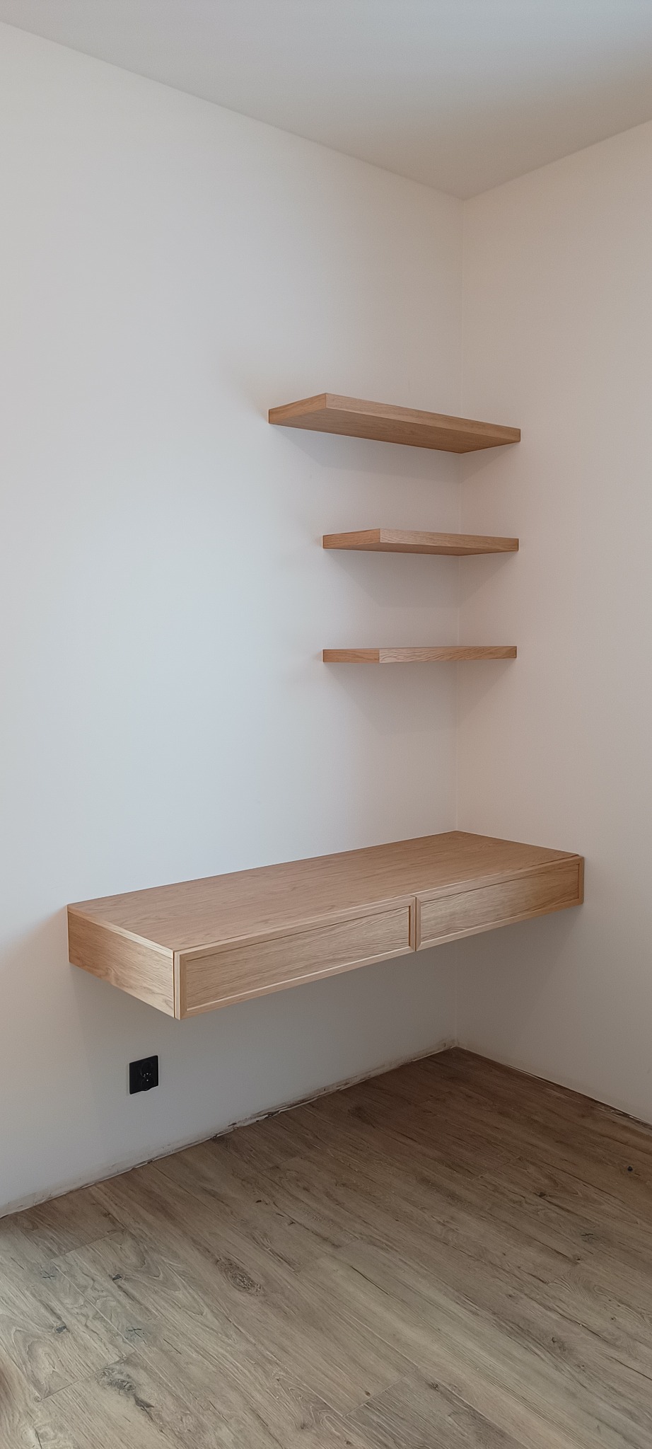 Jasne, drewniane półki wiszące i biurko z dwiema szufladami, zamontowane na ścianie w rogu pokoju z panelami podłogowymi. Minimalistyczny design.