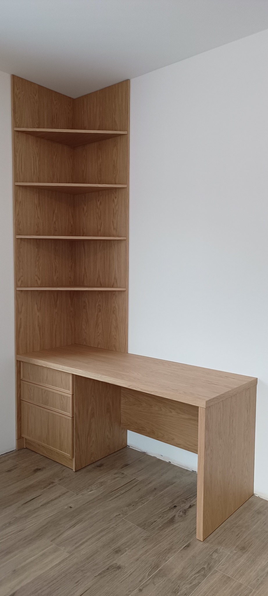 Kremowy, drewniany narożny regał z biurkiem i szufladami, dopasowany do jasnego wnętrza. Widoczne półki i minimalistyczny design.
