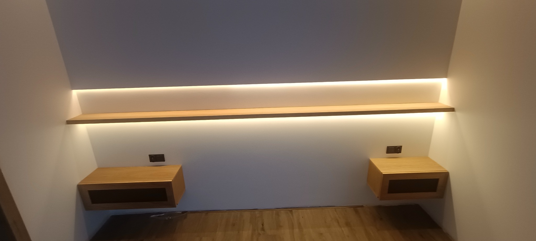 Drewniana szafka nocna z oświetleniem LED i półką, zamontowana na ścianie. Minimalistyczny design, ciepłe światło, widoczne gniazdka elektryczne. Wykończenie w naturalnym drewnie.