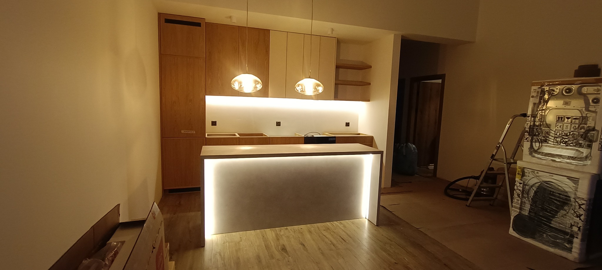 Nowoczesna kuchnia w trakcie montażu: drewniane szafki, podświetlany barek, wiszące lampy. Widoczne elementy wykończeniowe i narzędzia. Ciepłe oświetlenie.