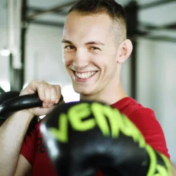 Uśmiechnięty mężczyzna w czerwonej koszulce trzymający kettlebell i rękawice bokserskie Venum z zielonym napisem, widoczne z bliska na tle siłowni.