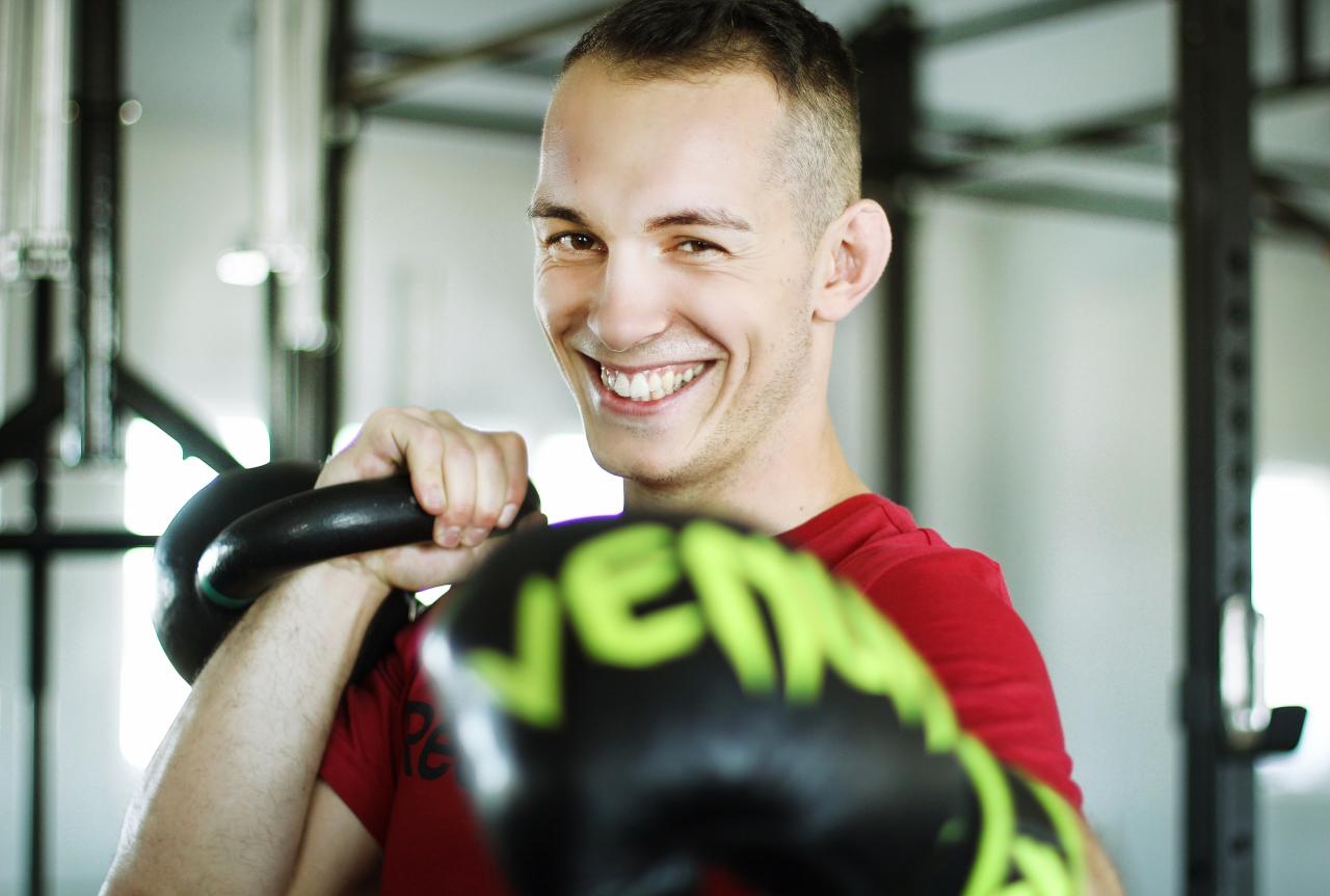 Uśmiechnięty mężczyzna w czerwonej koszulce trzymający kettlebell i rękawice bokserskie Venum z zielonym napisem, widoczne z bliska na tle siłowni.