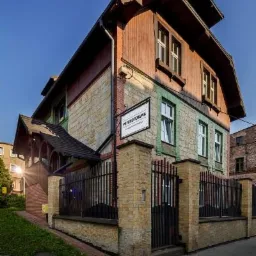 Elewacja kamienno-drewniana budynku z tabliczką informacyjną 'Psychoterapia' przy wejściu, otoczona czarnym metalowym ogrodzeniem.