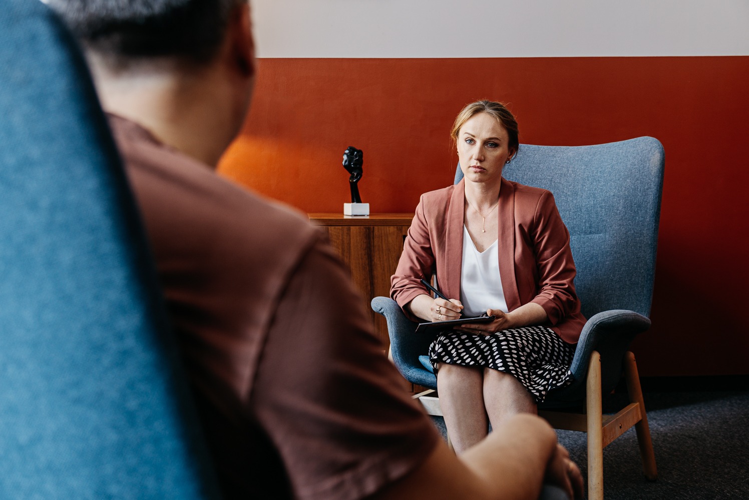 Kobieta w różowej marynarce siedzi na krześle, robi notatki podczas sesji. W tle pomarańczowa ściana i drewniana komoda z rzeźbą.