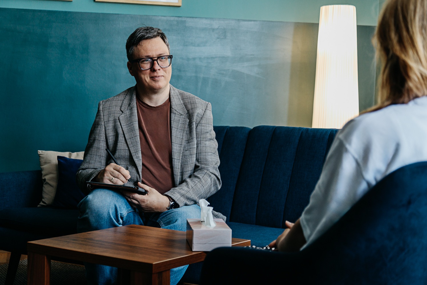 Sesja w Bytomiu: Mężczyzna w okularach siedzi na niebieskiej sofie, notując na tablecie. Pacjent siedzi naprzeciwko, widoczny od tyłu. Ciepłe oświetlenie, spokojna atmosfera.
