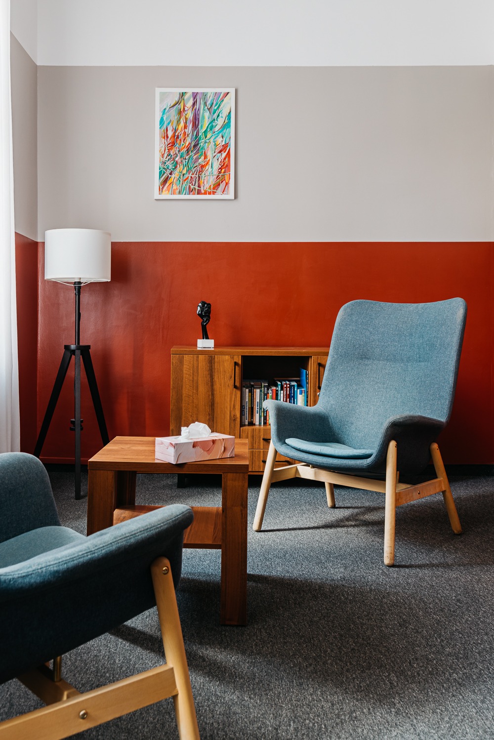 Wnętrze gabinetu z dwoma fotelami, stolikiem z chusteczkami, regałem z książkami i obrazem na ścianie w odcieniach szarości i czerwieni. Stonowana kolorystyka.