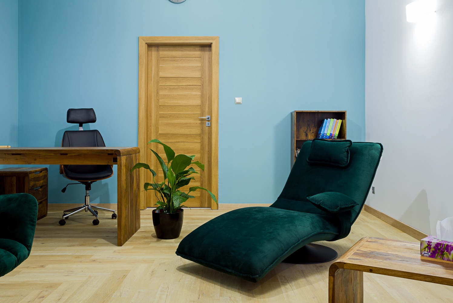 Gabinet z zielonym leżakiem relaksacyjnym, biurkiem z czarnym krzesłem i drewnianymi meblami na tle błękitnej ściany. Na biurku leżą książki.