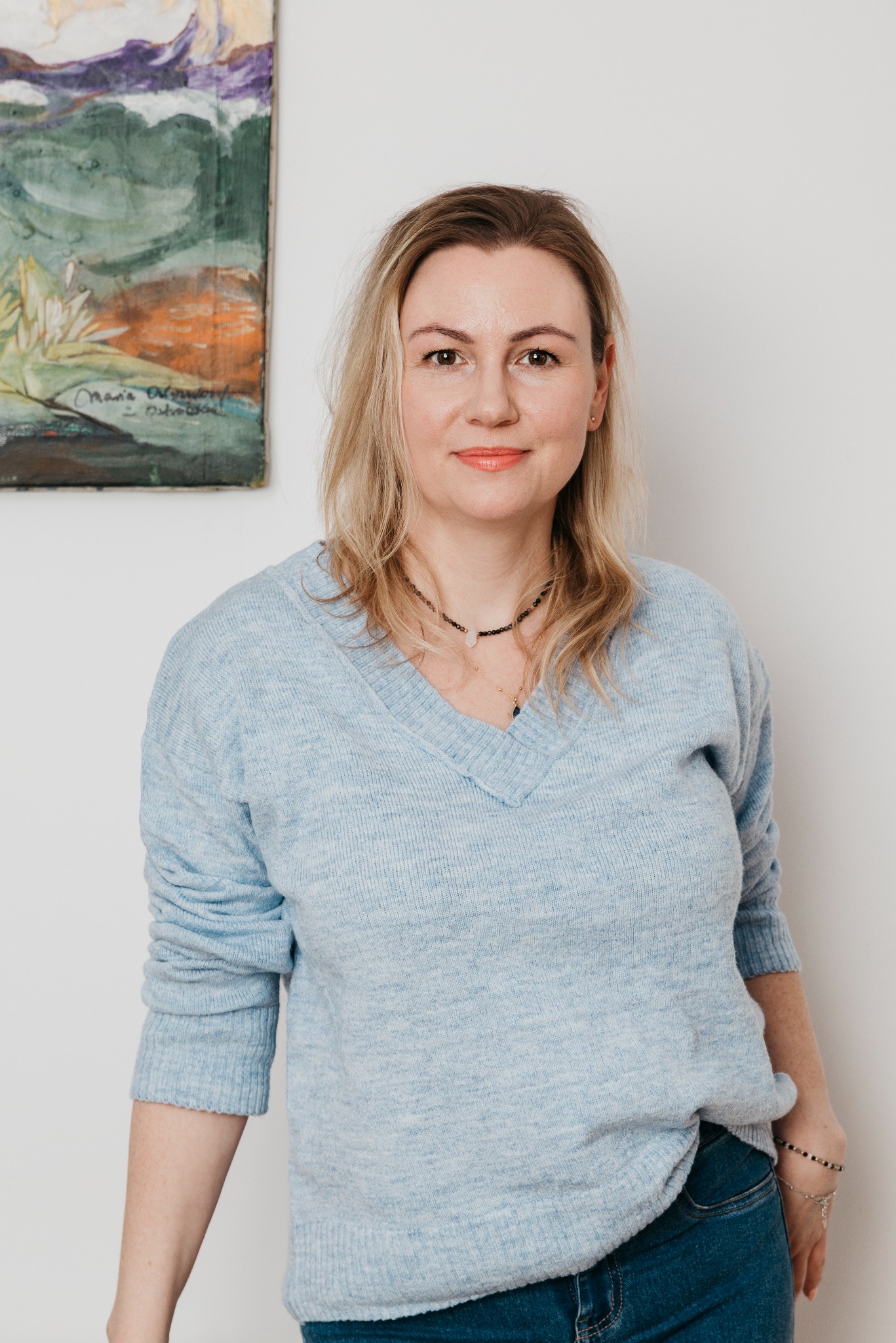 Portret kobiety w błękitnym swetrze na tle białej ściany z obrazem. Uśmiechnięta twarz, blond włosy, naszyjnik. Naturalne światło, neutralny wyraz twarzy.