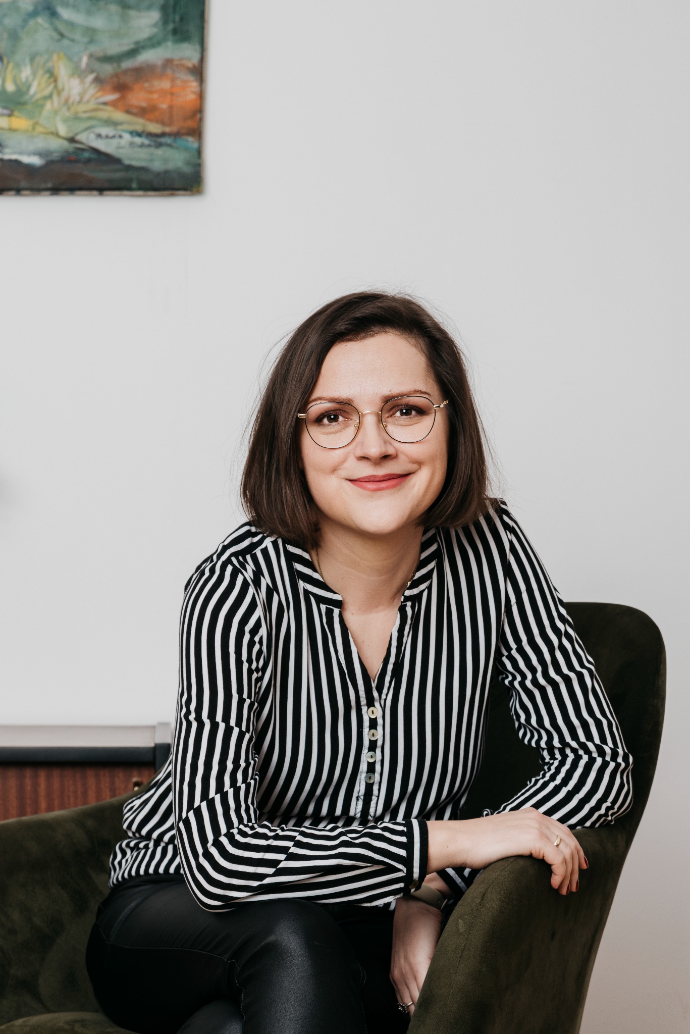 Portret uśmiechniętej kobiety w okularach, siedzącej w fotelu na tle białej ściany z obrazem. Ubrana w bluzkę w czarno-białe paski.