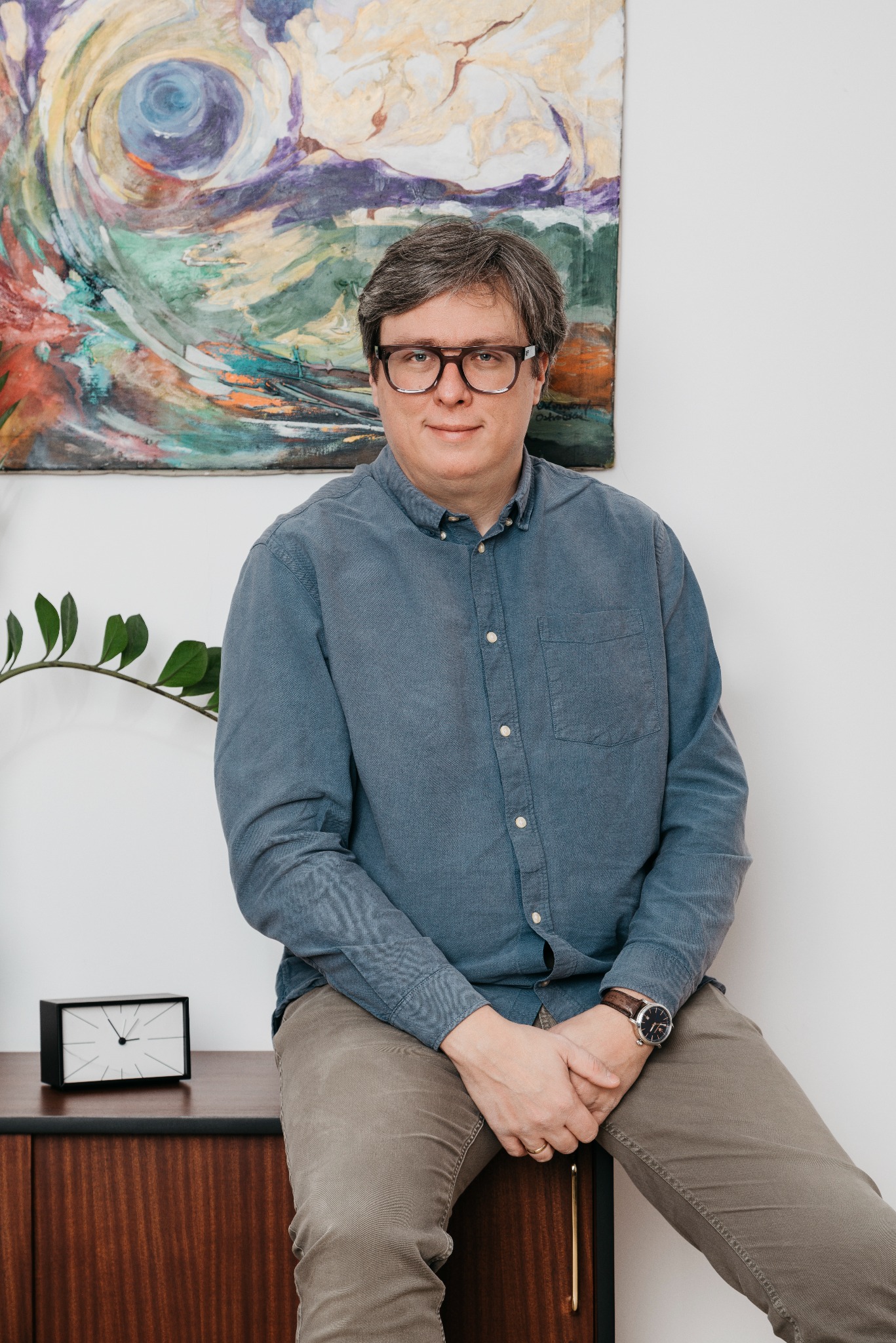 Portret mężczyzny w okularach siedzącego na szafce z zegarem, w tle abstrakcyjny obraz. Ubrany w niebieską koszulę i spodnie khaki. Naturalne światło, spokojna atmosfera.