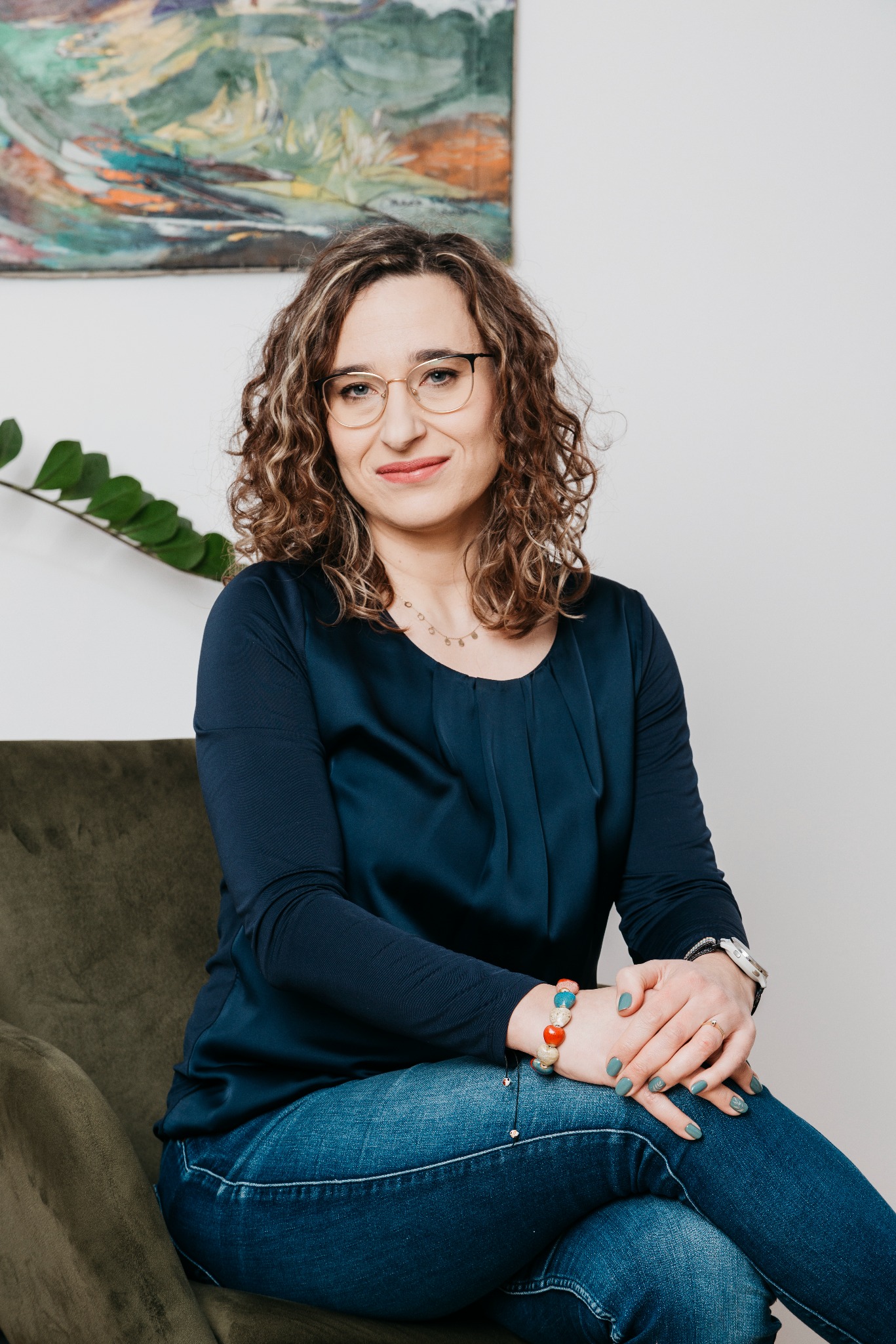 Kobieta w okularach siedzi na krześle, skrzyżowane nogi, dłonie splecione na kolanach. W tle obraz i roślina. Uśmiech, swobodna atmosfera.