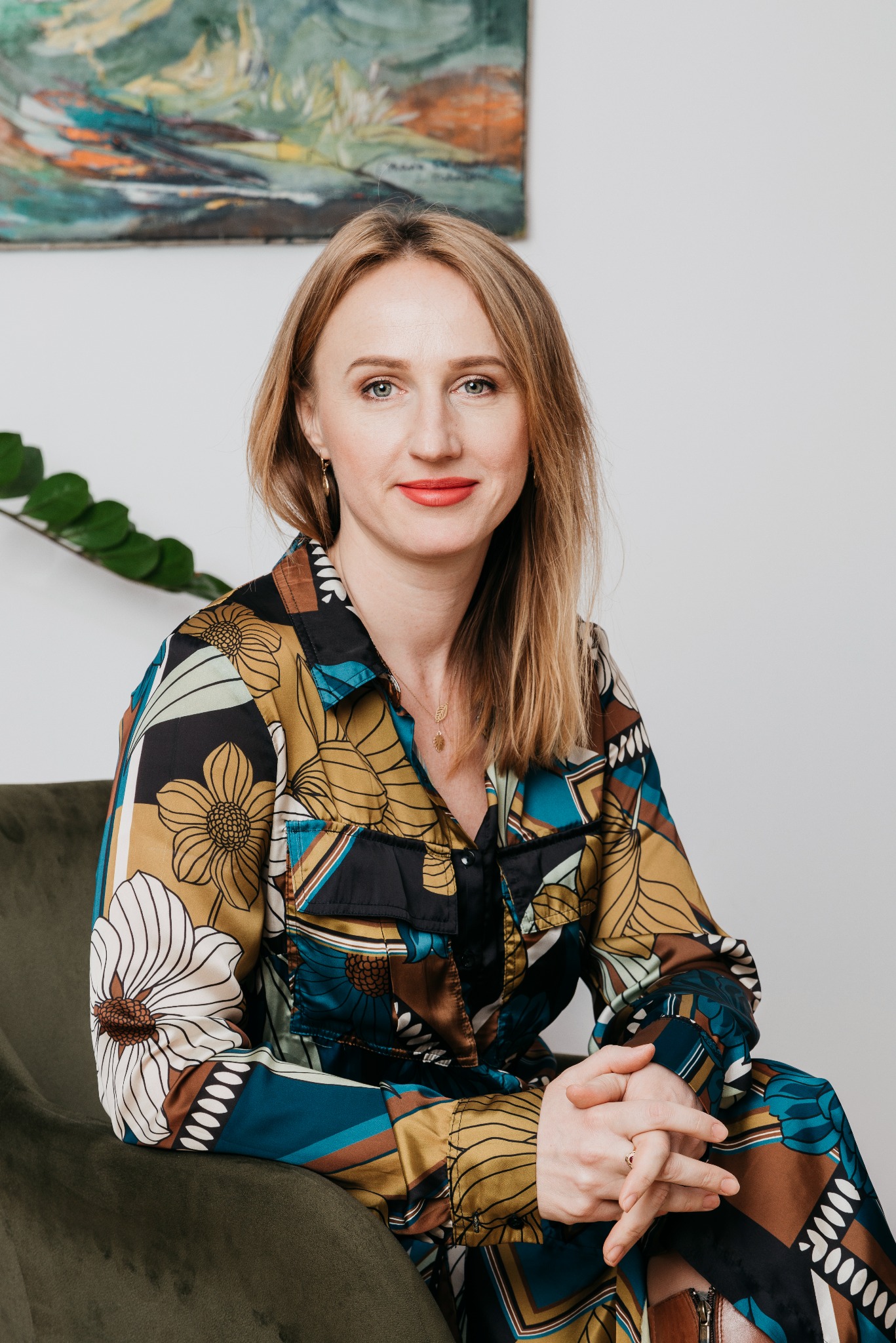 Portret kobiety z blond włosami, ubranej w wzorzystą koszulę, siedzącej na zielonym fotelu. W tle obraz i roślina. Uśmiech, kontakt wzrokowy, profesjonalny wygląd.