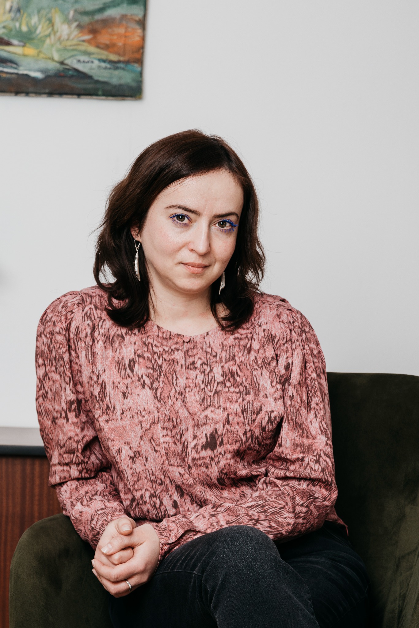 Portret kobiety z brązowymi włosami i niebieskim makijażem oczu, siedzącej na zielonym fotelu. W tle obraz na ścianie i drewniana boazeria.