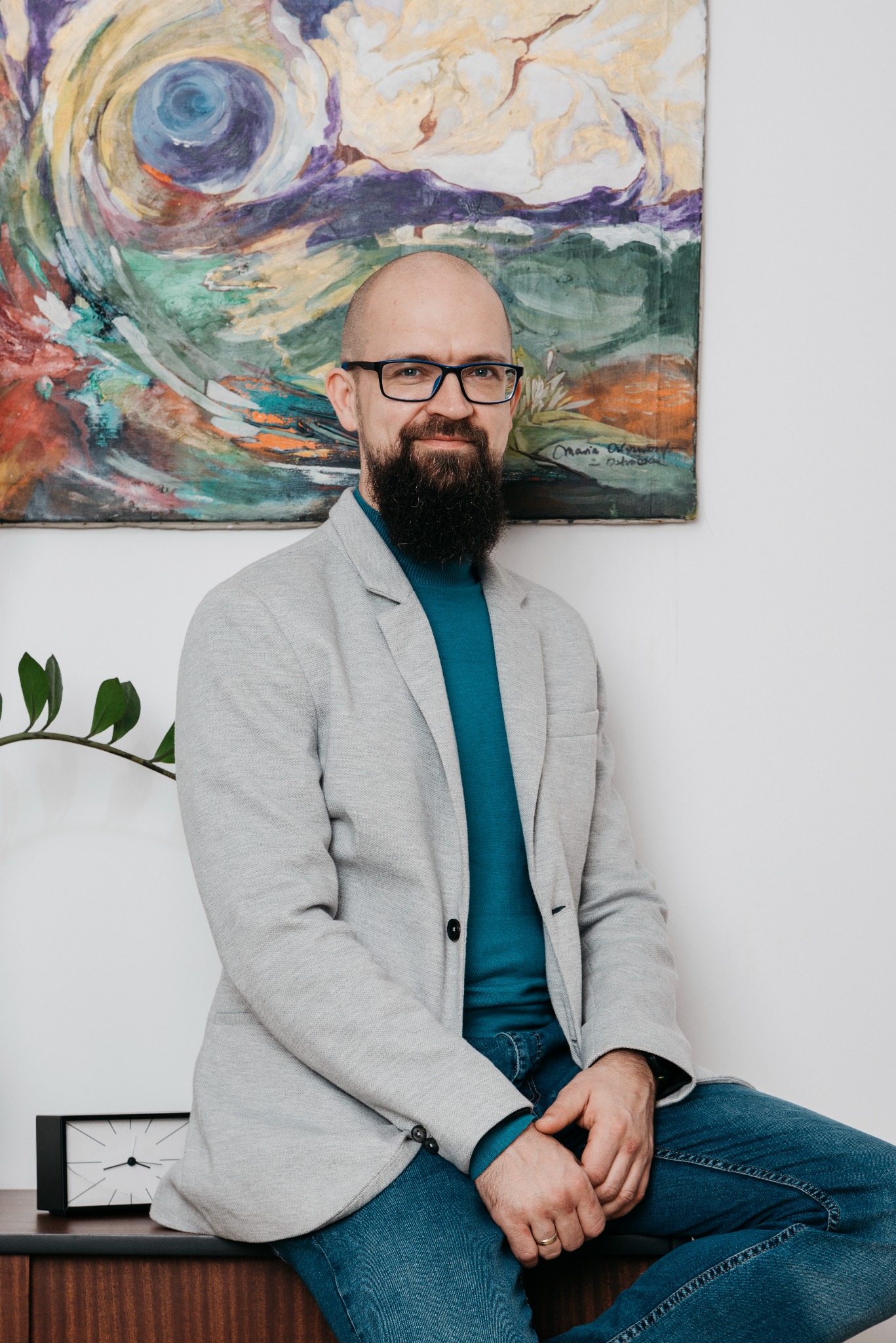 Portret mężczyzny z brodą w okularach, siedzącego na biurku, w tle abstrakcyjny obraz i roślina doniczkowa. Ubrany w szary blezer i niebieski golf.