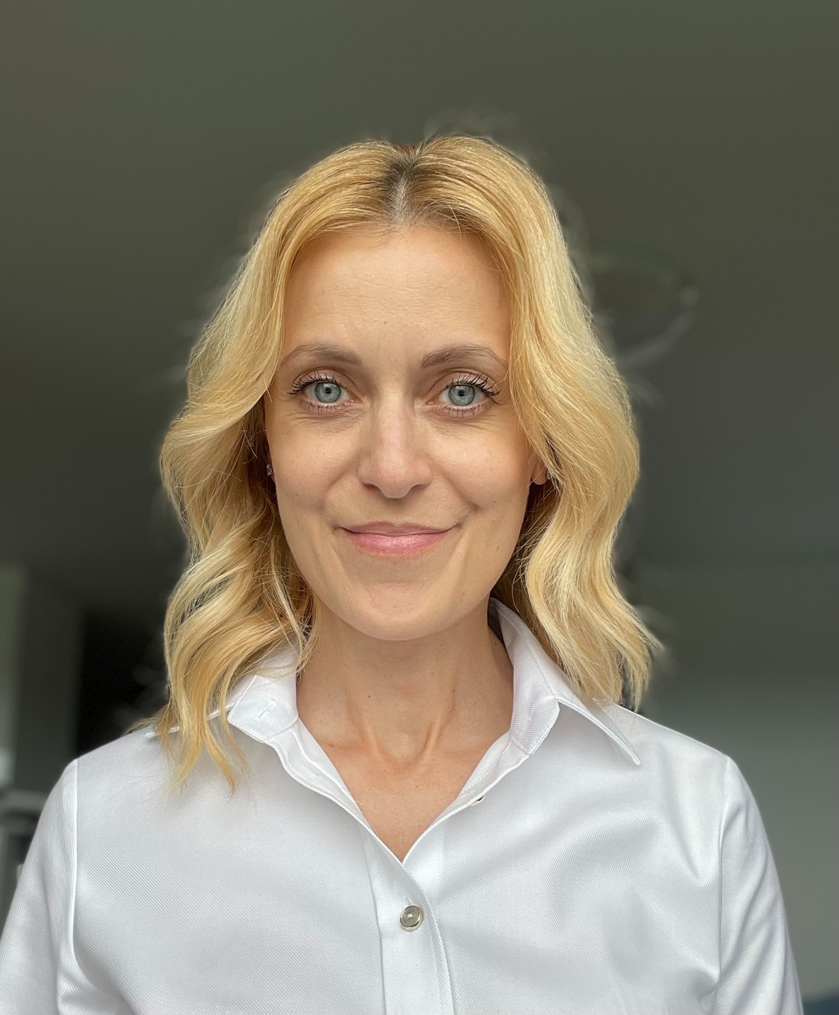 Portret uśmiechniętej blondynki o niebieskich oczach, w białej koszuli, na rozmytym tle. Profesjonalny wizerunek.