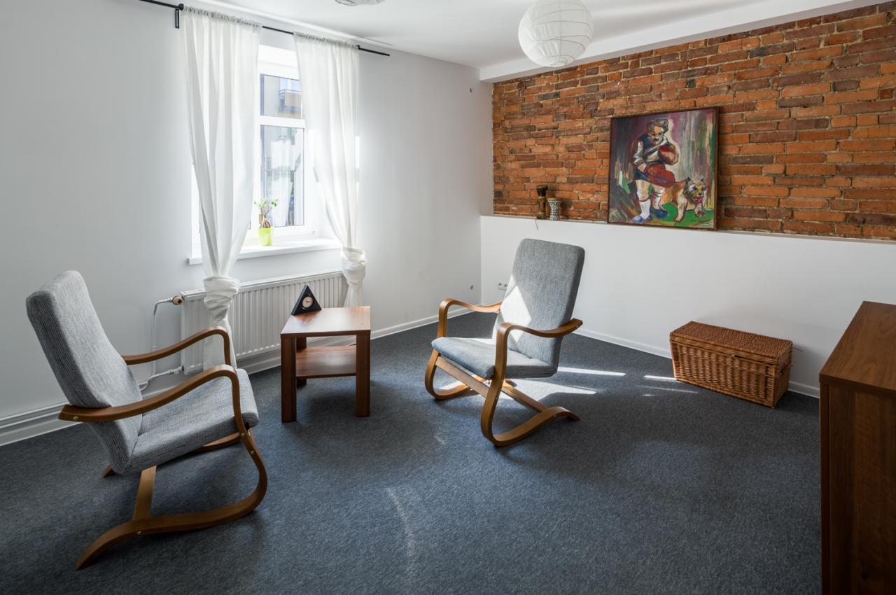 Jasny gabinet z dwoma fotelami bujanymi, małym stolikiem z zegarem, ścianą z cegły i obrazem chłopca z psem