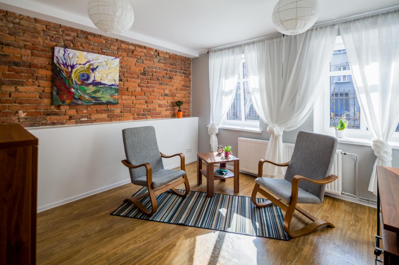 Jasne wnętrze gabinetu w Chorzowie z dwoma szarymi fotelami na drewnianych płozach, stolikiem kawowym z zegarem i książkami, ceglaną ścianą z abstrakcyjnym obrazem oraz oknami z białymi zasłonami.