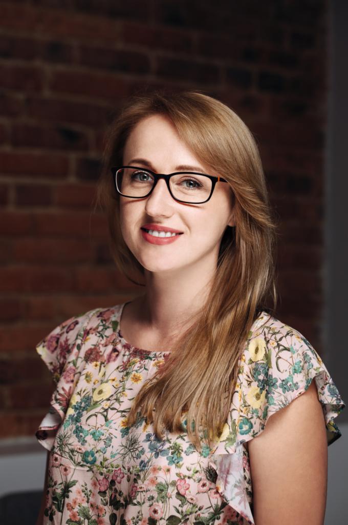 Portret uśmiechniętej kobiety w okularach i kwiecistej bluzce na tle ceglanej ściany.