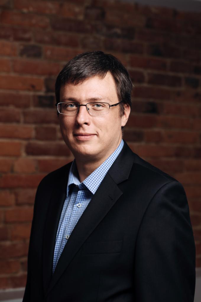 Portret mężczyzny w okularach i czarnym garniturze na tle ściany z czerwonej cegły.