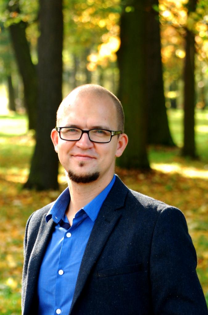 Portret mężczyzny w okularach, z kozim bródką, ubranego w niebieską koszulę i marynarkę, na tle jesiennego parku z opadłymi liśćmi.