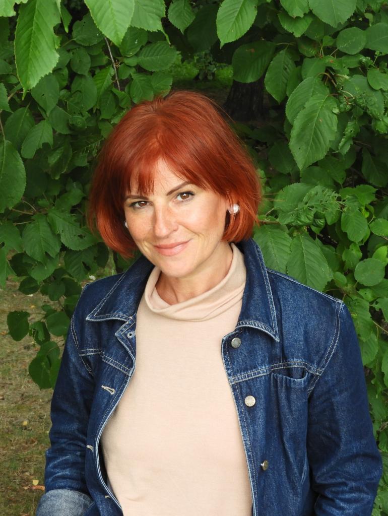 Portret uśmiechniętej kobiety z rudymi włosami w dżinsowej kurtce na tle zielonych liści.