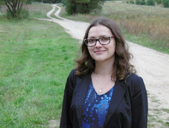 Uśmiechnięta kobieta w okularach i marynarce na tle polnej drogi.